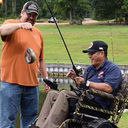 Disabled Access section link. Image depicts one person standing holding a fish that the other person in a track chair has just caught.