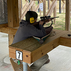Shooting Ranges link. Image depicts a person outdoors at a shooting range pointing a rifle at a target.